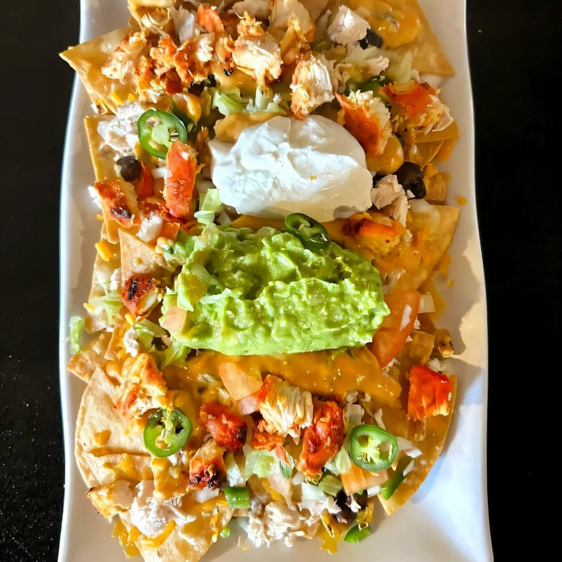 A plate of Fusion Tikka Taco nachos with chicken, cheese, guacamole, sour cream, and jalapeños.
